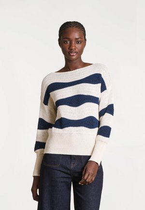 Femme noire aux cheveux tressés portant un pull en maille blanc avec des motifs de vagues bleu marine et un jean bleu foncé, debout devant un fond uni.