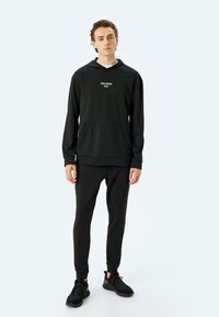Zwarte sportieve hoodie met voorzakken, met de tekst "FORCE STRENGTH FOCUS". Gecombineerd met bijpassende tapered sweatpants en zwarte sneakers.