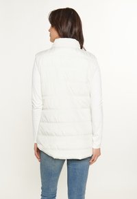 Weißer Puffy-Westen mit hohem Kragen, gestepptem Design und sichtbaren Nähten. Getragen über einem weißen Langarmshirt, kombiniert mit blauen Jeans.