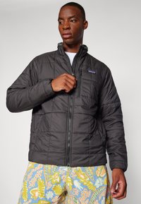 Veste matelassée noire avec un col haut, fermeture éclair à l'avant et petite poche poitrine. Le mannequin porte un short à motifs colorés.
