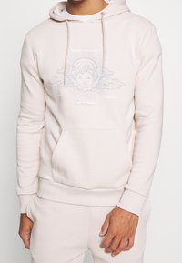 Felpa rosa chiaro in tessuto morbido con tasca frontale. Presenta una grafica di un cherubino con ali, circondato da linee di raggi solari.