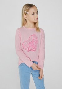 Růžová košile s dlouhým rukávem, která má na sobě grafiku postavy s dlouhými vlasy a volánkovými detaily, v kombinaci se svetle modrými džíny.