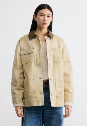 Nooruk, kellel on pikk tumedat värvi juuksed, kannab beežikat jopet nelja taskuga, mille all on kootud särk ja sinised teksad, seisab valge tausta ees.