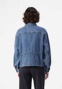 Veste en denim à coupe décontractée, de couleur bleue, avec une taille élastique et des manches retroussées. Présente un empiècement au dos et un minimum de détails.