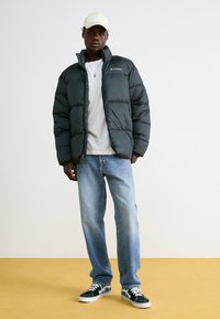 Sort pufferjakke med quiltet tekstur, lynlås, Columbia-logo; parret med lyseblå jeans og mørke sneakers, sat mod en neutral baggrund.