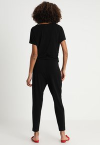 Zwarte jumpsuit met korte mouwen, ontspannen pasvorm, met een rits aan de achterkant, taps toelopende broekspijpen en een gladde stof. Rode sandalen accentueren de look.