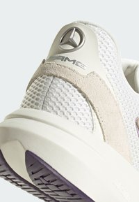 Białe buty sportowe z siateczkową cholewką i skórzanymi akcentami, z metalicznym logo oraz teksturowaną podeszwą z purpurowym akcentem.