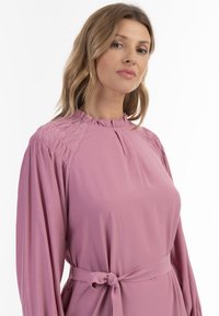 Šaty s dlouhým rukávem z jemné růžové tkaniny, které mají na ramenou nařasený design, vysoký výstřih a zavázaný pas pro přiléhavý střih.