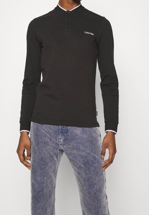 Homme portant une chemise noire à manches longues Calvin Klein avec des boutons au col et un pantalon en velours côtelé bleu clair.