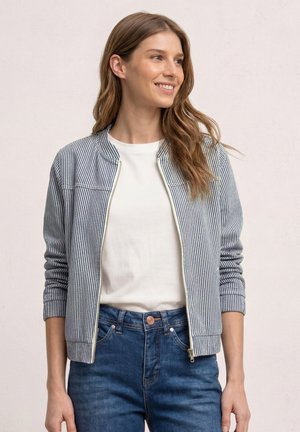 Smilende kvinde iført hvid skjorte, blå- og hvidstribet lynlåstrøje og blå jeans, stående foran en ensfarvet lys baggrund.