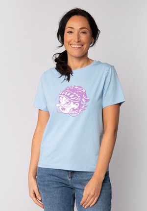 Frau lächelt, trägt ein hellblaues T-Shirt mit Grafik eines lila Comicgesichts und Creolen, kombiniert mit blauen Jeans, steht vor einem schlichten Hintergrund.