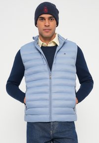 Világoskék pufi mellény függőleges varrással, teljes cipzárral és oldalzsebekkel; sötétkék pulóver és világos színű ing fölött viselve.