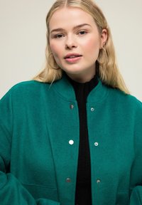 Tyrkysová vlněná bunda s vysokým límcem a cvočkovými knoflíky, navrstvená přes černý rolák. Modelka má velké kruhové náušnice.