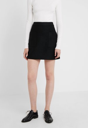 Kobieta w czarnej mini spódnicy z przednimi naszywanymi kieszeniami, białej bluzce z długim rękawem i czarnych butach na sznurowadła, stojąca na białej podłodze.