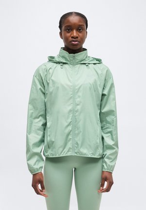 Žena v svetlozelenej mikine s kapucňou a na zapínanie na zips a v zodpovedajúcich legínach, stojaca čelom dopredu s neutrálnym výrazom.