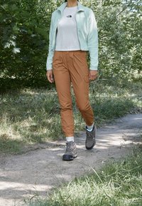 Giacca zip-up azzurro chiaro sopra una camicia bianca, pantaloni cargo marroni e stivali da escursione grigi. Lo sfondo presenta fogliame verde e un sentiero di terra.