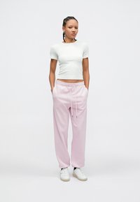 Λευκό στενό T-shirt σε συνδυασμό με ανοιχτό ροζ φόρμα με ελαστική μέση και κορδόνι. Το μοντέλο φοράει λευκά αθλητικά παπούτσια.