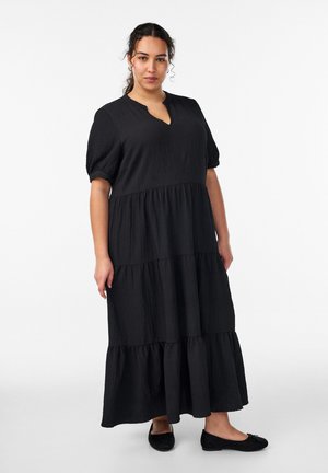 Kobieta stojąca, ubrana w długą czarną sukienkę wielowarstwową z krótkimi bufiastymi rękawami i czarne baletki na białym tle.