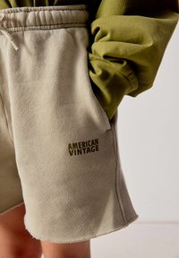 Beige sweatshorts met een trekkoord in de taille en het logo "AMERICAN VINTAGE", gedragen met een olijfgroene top met lange mouwen, hand in de zak.