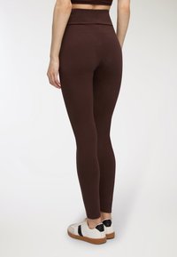 Leggings marroni a vita alta realizzati in un morbido tessuto elasticizzato. Presentano una silhouette aderente e cuciture laterali, abbinati a sneakers bianche e nere.