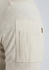 Beige Pullover mit strukturiertem Strickdesign, Rippdetails und einer quadratischen Stoff-Patchtasche am oberen Arm.