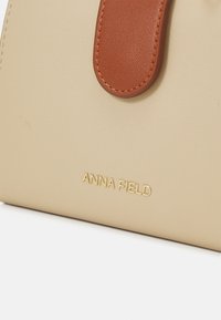 Borsa a mano in pelle sintetica beige con una tracolla in pelle marrone e logo "ANNA FIELD" in oro stampato sul davanti. Texture liscia con dettagli minimi.