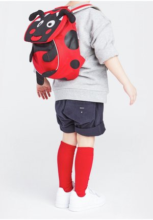 Sac à dos sur le thème de la coccinelle rouge avec des taches noires, deux antennes et un visage souriant. Porté par un enfant en sweat gris, shorts bleu marine et chaussettes rouges.