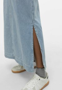 Črtasta denim maksi krilo s stranskimi razpoki, svetlo modre barve, z razmršenim robom, v kombinaciji z belimi supergami in sivimi nogavicami.