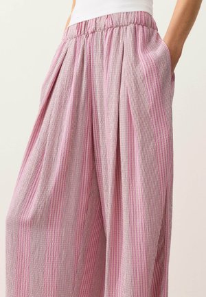 Losse roze gestreepte broek met plooien en een elastische tailleband, gedragen met een wit shirt, handen in de zakken.