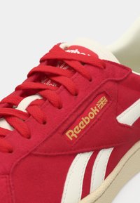 Sneaker rossa in suede con accenti in pelle bianca, dotata di chiusura con lacci, superficie testurizzata e logo Reebok dorato sul lato.