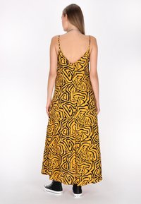 Rochie maxi galbenă cu un model abstract negru, design cu spate în V, curele subțiri și o siluetă fluidă, asociată cu adidași negri înalți.