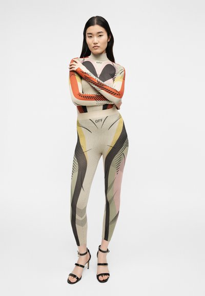 Γυναίκα όρθια με σταυρωμένα χέρια, φοράει έναν πολύχρωμο, με σχέδια, εφαρμοστό ολόσωμο φόρεμα και μαύρα σανδάλια με ψηλά τακούνια σε απλό φόντο.