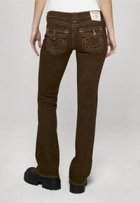 Hnědé rozšířené džíny s kontrastním bílým šitím, dvěma zadními kapsami s ozdobnou výšivkou a značkovou štítkem na pasu.