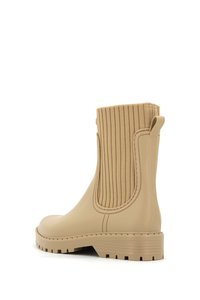 Bota impermeable beige con un panel elástico texturizado y acanalado, punta redondeada y diseño de suela dentada. Cuenta con una lengüeta para facilitar su puesta.