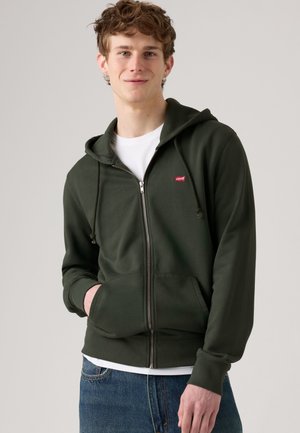 Młody mężczyzna noszący ciemnozieloną bluzę z kapturem i zamkiem błyskawicznym z logo Levi's, białą koszulkę i niebieskie jeansy, uśmiechający się z jedną ręką w kieszeni.