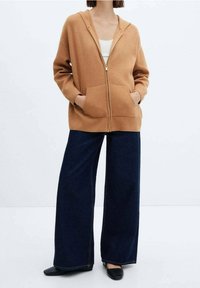 Sudadera marrón con cremallera hecha de tela suave, con un bolsillo frontal, combinada con pantalones de mezclilla oscuros de pierna ancha y zapatos planos negros.