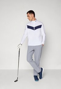 Top de golfe branco de manga comprida com design em chevron azul-marinho; calças cinzentas; em pé numa pose descontraída, segurando um taco de golfe, usando uma luva e sapatos.