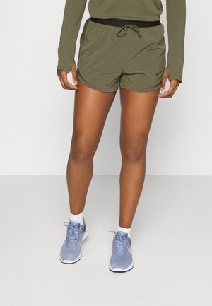 Legs wearing olive green athletic shorts, long-sleeve top, white socks, and light blue running shoes standing on a plain light gray floor.