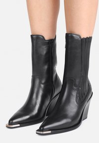 Stivali wedge in pelle nera a metà polpaccio con punta affusolata, cerniera laterale e coperture metalliche sulla punta indossati su gambe nude su sfondo bianco.