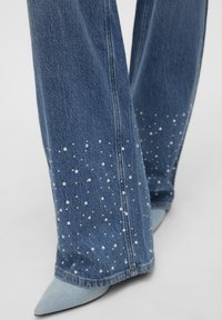 Jeans a zampa d'elefante in denim con decorazioni in perle all'orlo, caratterizzati da un colore azzurro chiaro e una leggera variazione di texture nella parte inferiore.