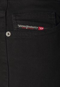 Svart denimjeans har en framficka med en röd och vit tygetikett som säger "Diesel Industry Denim Division", fäst med metallhardware.