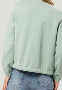 Woman wearing light green long-sleeve top with elastic cuffs and hem paired with blue denim jeans, shown from the back.