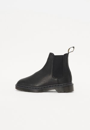 Schwarzer Chelsea-Stiefel aus Leder mit elastischem Seiteneinsatz, Zuglasche an der Ferse und dicker Gummisohle, seitlich vor weißem Hintergrund dargestellt.