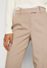 Beige tailored trousers with a smooth texture, featuring a matching belt and side pockets, paired with a cream long-sleeve shirt.
