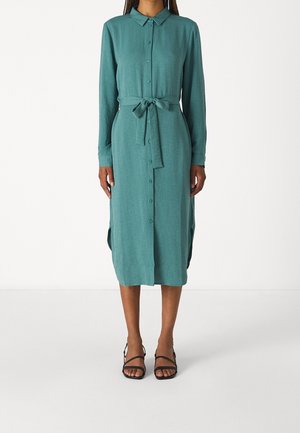 Frau trägt ein tealfarbenes, langärmliges Midi-Kleid mit Knöpfen, einem gebundenen Taillengürtel und schwarzen offenen Sandalen, steht auf einem weißen Boden.