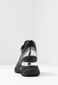 Chaussure de sport noire et grise avec une tige texturée, un accent métallique au talon et une semelle épaisse en caoutchouc noir.