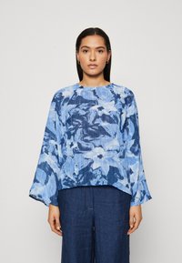Blusa blu a fantasia con maniche lunghe e scollatura rotonda, realizzata in un tessuto leggero e fluido con una vestibilità morbida. Abbinata a pantaloni in denim scuro.