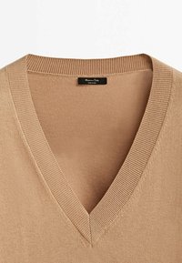 Pull beige col en V avec col côtelé, en tissu tricoté fin, avec une petite étiquette noire à l'arrière du cou.
