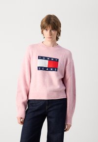 Šviesiai rožinė megzta džemperis su raštuotomis rankovėmis ir apačia, kuriame yra tamsiai mėlynas, raudonas ir baltas logotipo paminkštinimas su užrašu "TOMMY JEANS."