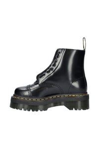 Bottine en cuir noir avec une semelle à crampons épaisse, présentant un laçage décoratif, un empiècement cousu jaune et une languette au niveau du talon.